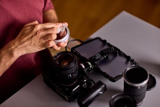 Focused young man cleaning camera lens with white microfiber cloth indoors in warm low light studio, holding lens in left hand closeup portrait side view scene