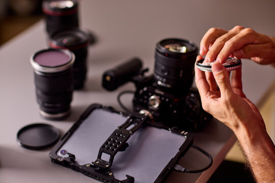 Focused young man cleaning camera lens with white microfiber cloth indoors in warm low light studio, holding lens in left hand closeup portrait side view scene