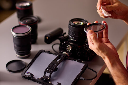 Focused young man cleaning camera lens with white microfiber cloth indoors in warm low light studio, holding lens in left hand closeup portrait side view scene