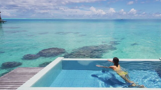 Summer sunbathing happy bikini woman swimming in infinity pool of luxury resort hotel relaxing in idyllic overwater ocean view suite. Suntan solar skin care. Raw unedited video of real woman model.