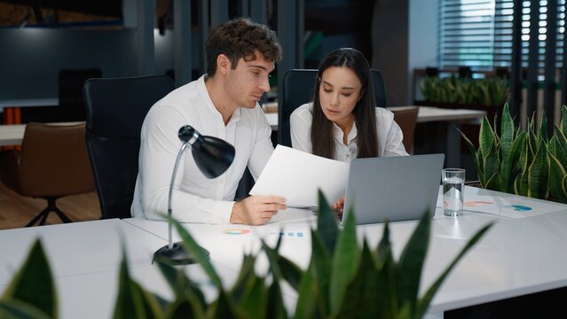 Two business partners talking looking in papers with stats data graphs of project marketing result reading documents computer work in office businesswoman with businessman talk working look in laptop