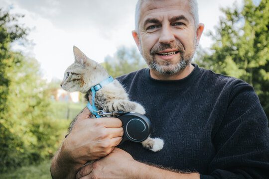 Middle-aged man with gray beard holds a tabby cat wearing a blue harness while standing outdoors in a green park with trees in the background