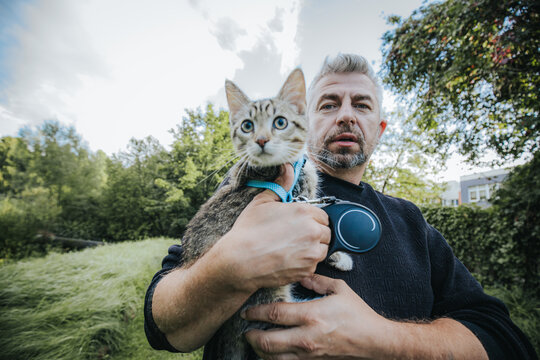 Man holding a tabby cat outdoors in a green field with trees and cloudy sky in the background, showcasing a close-up view of both subjects