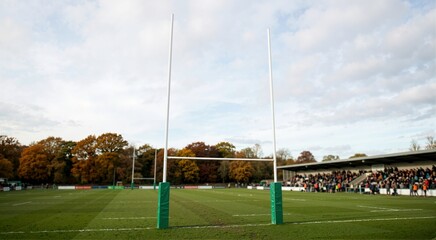 Fototapeta premium Rugby goal posts on a green grass field. Stadium stand with spectators and autumn trees under cloudy sky