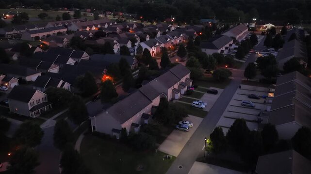Townhome rooftops spread through dusky neighborhood as windows glow warmly, parked cars line quiet driveways, tall pine trees shade winding streets throughout suburb. Aerial drone flyover shot
