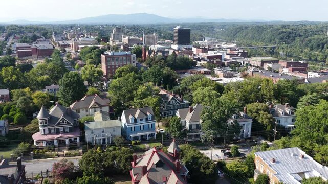 Victorian rooftops fill tree lined Lynchburg neighborhood, downtown high rises and church spires rise beyond dense canopy, distant mountains fade into hazy summer sky. Aerial drone flyover shot