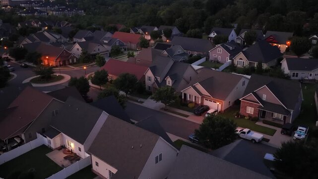 Suburban rooftops glow beneath dusk sky as porch lights flicker on, curved driveways lead toward garage doors, mature trees shade quiet cul de sac neighborhood streets. Aerial drone flyover shot