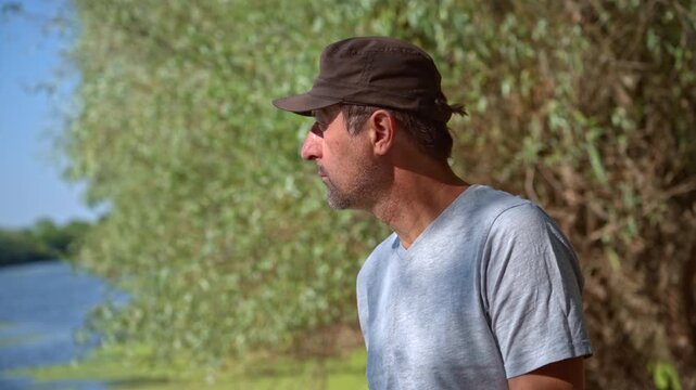Profile of a man in a cap looking over a river with lush green trees in bright daylight. 4K.