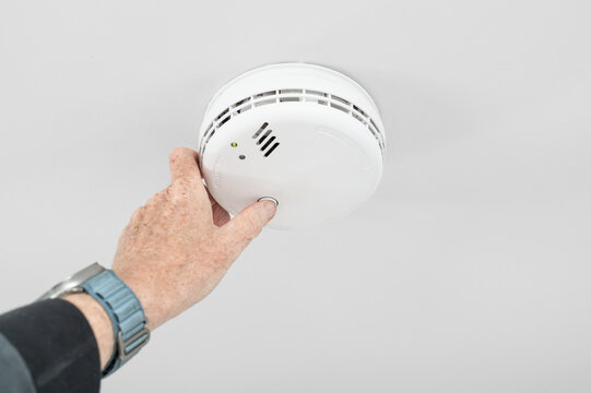Handyman seen doing a weekly test of a combine smoke and carbon monoxide tester in a home hallway. The detector is wired into the house wiring a requires no battery.