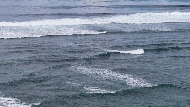 Aerial view of rolling ocean waves with white surf and textured blue-gray water under an overcast sky, conveying calm, moody seascape energy and natural coastal motion and patterns