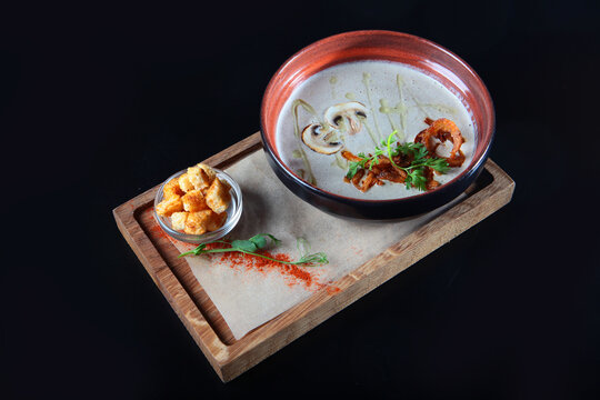Creamy mushroom soup in ceramic bowl. Garnished with parsley, oil, pepper. Croutons in small glass bowl. Wooden board. Healthy meal. Dark background.