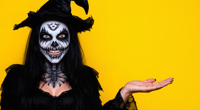 Frightening woman wearing black costume and halloween makeup holding copyspace on palm, isolated over yellow background
