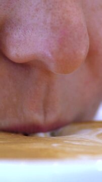 Close up adult man sips coffee from cup, lips and nose near creamy foam. Man drinks calmly in macro style, with focused expression and tight crop on face, showing mouth, chin, and white ceramic cup.