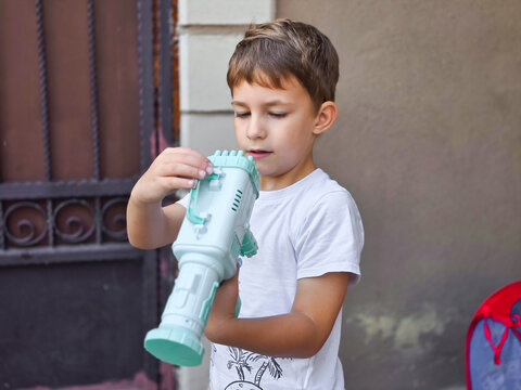 Boy playing with bubble gun outdoor, happy child blowing soap bubbles in summer, kid having fun with bubble blaster toy near house