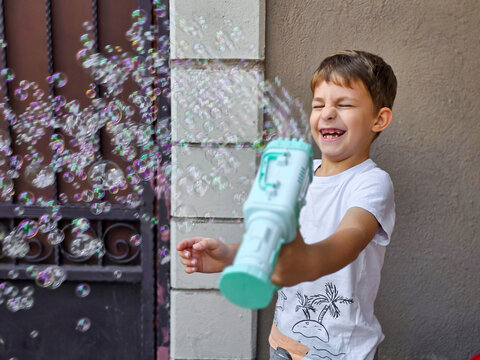 Boy playing with bubble gun outdoor, happy child blowing soap bubbles in summer, kid having fun with bubble blaster toy near house