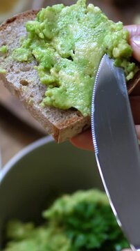 Hand spreads avocado mash onto bread slice with knife, showing concentrated kitchen prep and rustic style. Bread slice and green topping fill frame while smooth spreading continues.