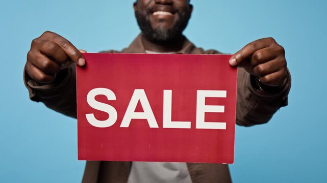 Midsection shot of African American man holding red sale sign against blue background. Ideal for retail promotion, seasonal discount marketing, and ecommerce shopping campaigns