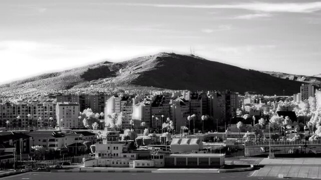 Infrared timelapse of the Gibraltar airport and cityscape below Sierra Carbonera mountain as shadows shift and clouds pass across the sky