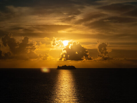 A cruise ship glides in silhouette across the Caribbean Sea as the sun sets behind dramatic golden clouds, casting a warm reflection across the calm water.