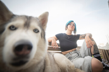 Trendy gen z woman with blue hair resting on bench with blurred husky on foreground with copy space © Aliaksei