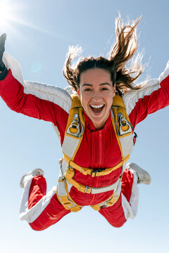 A thrilled young woman smiling and screaming with joy while skydiving in free fall, wearing a red and white jumpsuit with a yellow harness against a clear blue sky background.