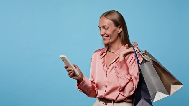 Caucasian woman holding shopping bags looking at smartphone with excited expression. Clip is ideal for ecommerce marketing, digital sales promos and retail campaigns