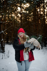 Fototapeta premium Smiling young woman in winter outfit holding pine branches outdoors in snowy forest. Concept of Christmas holidays, winter lifestyle, happiness, seasonal nature and festive mood.