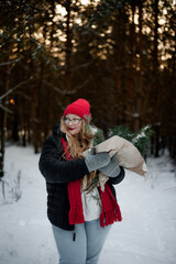 Fototapeta premium Smiling young woman in winter outfit holding pine branches outdoors in snowy forest. Concept of Christmas holidays, winter lifestyle, happiness, seasonal nature and festive mood.