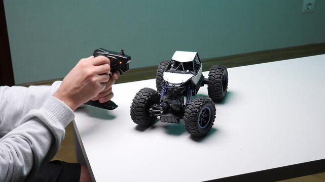 Person's hands holding a black radio remote control, manipulating a white and black toy rock crawler car with large tires sitting on a white table, engaging in hobby and leisure activity indoors