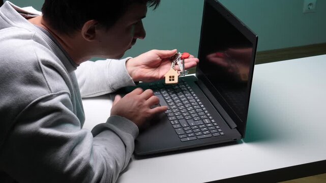 Young man holding a set of house keys with a wooden house shaped keychain while typing on a laptop, searching for real estate, property, or rental options online from home