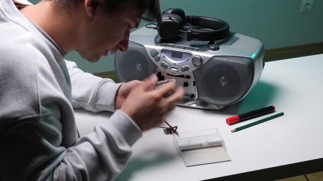 young man holding a broken audio cassette tape with tangled magnetic film, feeling frustrated after experiencing an issue with vintage technology and an old boombox stereo system