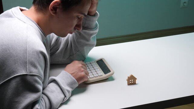 Man feeling stressed and overwhelmed, covering his face while calculating mortgage payments and potential debt with a calculator next to a small house model