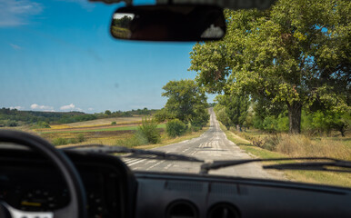 Obraz premium View through a car windshield of a rural country road in Romania, lined with trees and bordered by farmland, stretching toward distant hills under a clear blue sky.