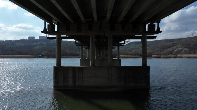 Moving beneath a concrete bridge over a calm river, passing structural piers and sunlit water. Transport infrastructure, engineering, river crossing
