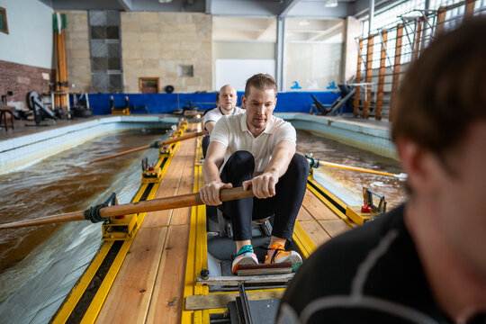 Focused male rower reaches catch position while teammates continue training behind. Tense expression and body posture accompany strength, endurance, determination and intense physical fatigue.