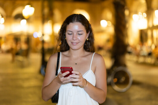 Young woman with a mobile phone in her hands near night bars and restaurants, she answers messages to friends and calls a taxi on the app.