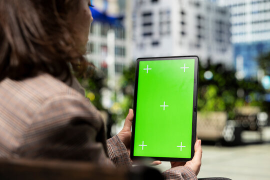 Woman reads a digital book on a green screen tablet during a sunny city morning, relaxing on a park bench with a chroma key ebook reader in close up for outdoor entertainment and digital leisure.