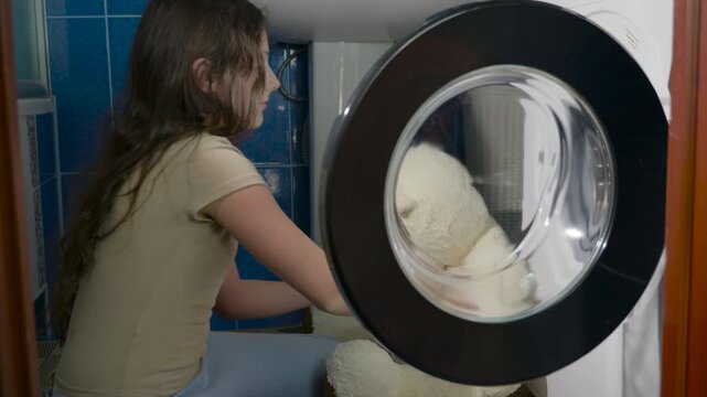 Young girl putting large teddy bear in washing machine. Careful young girl tenderly placing her large white stuffed teddy bear into the washing machine drum to clean it, demonstrating responsibility