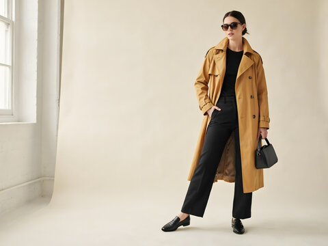 A woman wearing a tan trench coat, black trousers, and sunglasses, holding a black handbag while standing against a light beige studio background