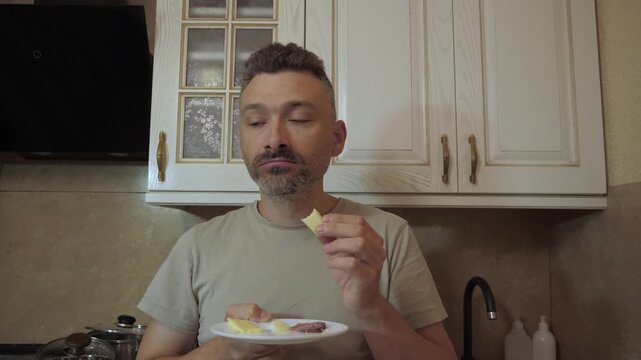 Man holds plate of sliced cheese and sausage while lifting bite sized food to mouth and chewing, with calm focused expression and slight enjoyment, medium frontal view shows upper body
