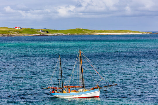Two-masted ketch resting on turquoise sea water with furled red sails and rigging, cottages along a grassy shore visible in the distance