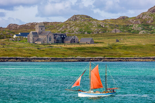 Traditional sailboat with red sails navigating coastal water past ancient Iona Abbey, grassy hills and rocky coastline behind, Inner Hebrides, Scotland