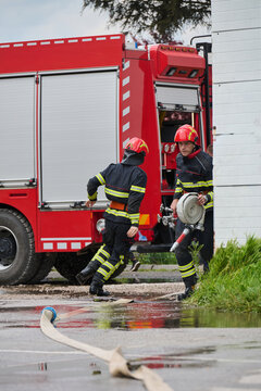 Two Firefighters Unrolling Hose Beside Red Fire Truck During Emergency Response At Scene