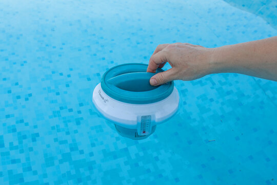 Hand placing a chlorine tablet into a floating dispenser with an integrated thermometer, ensuring proper water treatment and hygiene for a clean and refreshing swimming pool during summer