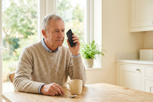 A grey-haired man in his sixties holds a phone away from his ear with a thoughtful, uncertain expression at a sunlit kitchen table. Voice cloning scam awareness and elder fraud prevention concept.
