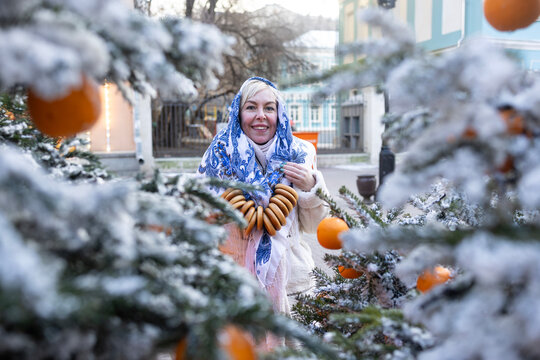 Smiling woman in traditional Russian shawl holding string bag with mandarins and sushki by snowy Christmas tree