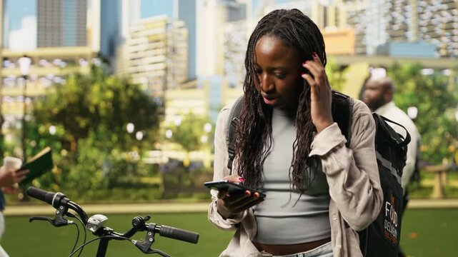 Courier fidgets and checks a food delivery app for a new order, waiting anxiously for a request to work in the gig economy at a city park.