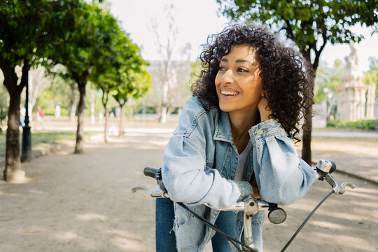 Young adult happy woman with curly hair with a bicycle smiling softly and touching her hair outdoors in the city. She is looking away. Friendship and travel concept.