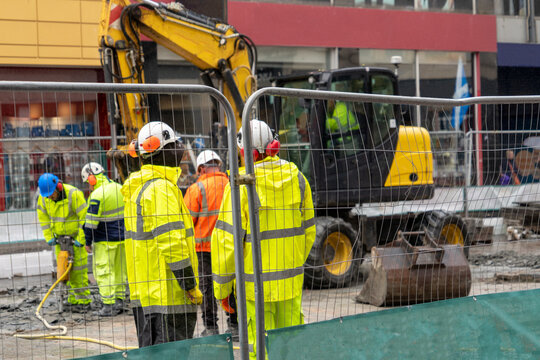 Construction workers on an outdoor urban work site, cordoned off by a metal wire mesh fence. Wearing high-visibility safety gear and hard hats at an active street excavation site with an excavator. 
