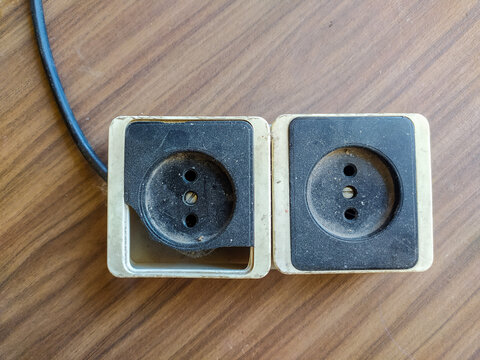 This vintage, dusty double electrical socket lies on a worn wooden table. Evoking nostalgia, obsolescence, and neglect, this broken plastic power outlet has nobody around.